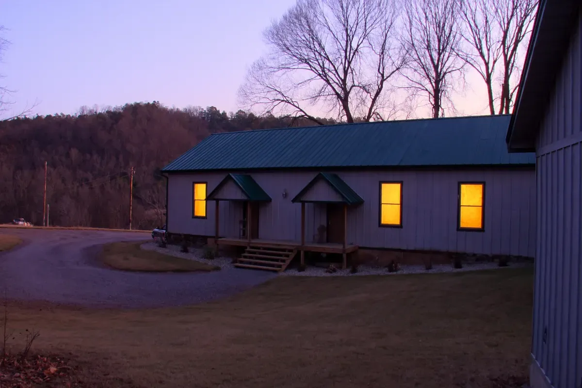 Modest finished post-frame home at dusk with lit windows and a gravel drive in rural Alabama